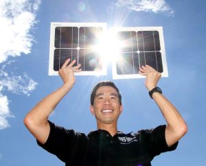 A man smiles and holds a solar panel above his head with the sun shining through the middle.