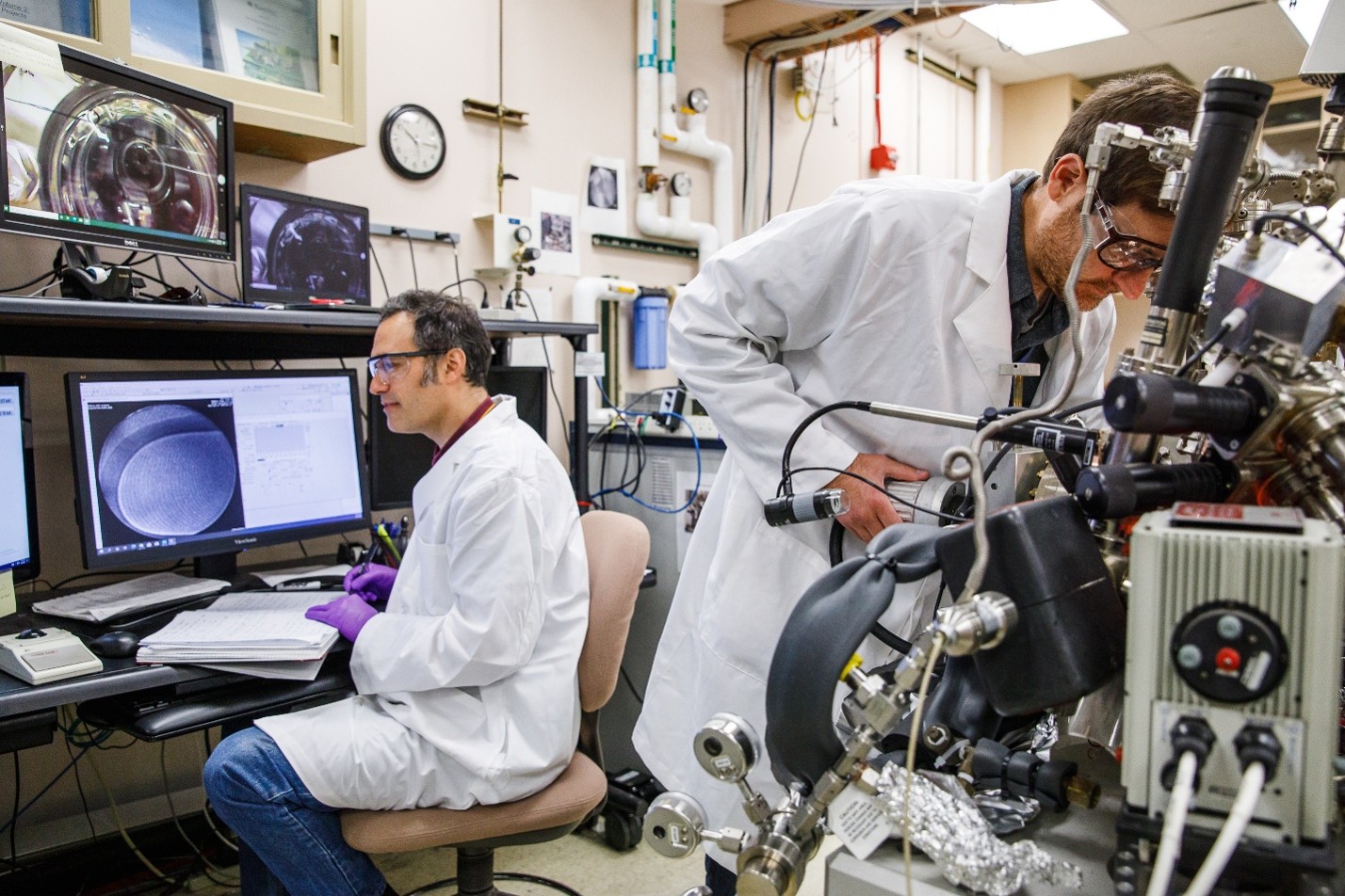 A man in a white lab coat and safety glasses sits at a computer while writing in a notebook. Another man in a white lab coat and safety glasses works on a lab instrument.