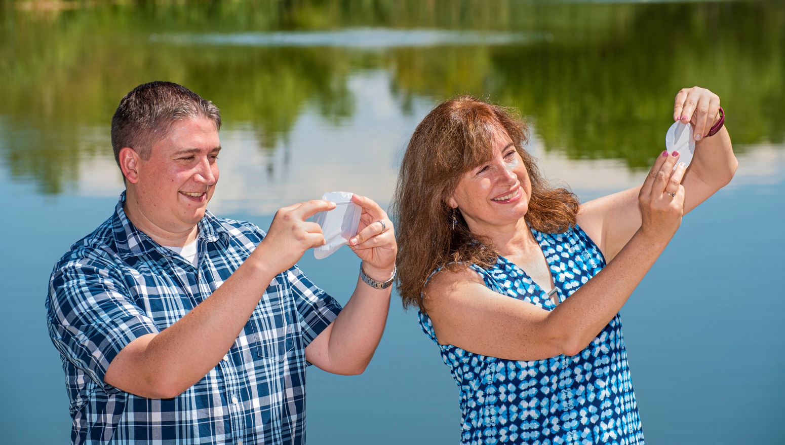 Two scientists look at hand-sized white membranes, water and lush trees in background.