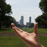 A hand hold a circular piece of Aerigel, which you can see park and city skyline through.