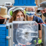 Technicians and students examine a plexiglass cube fractured by a small explosion