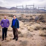 Two researchers stand in the desert. Power transformers are on the horizon in the background.