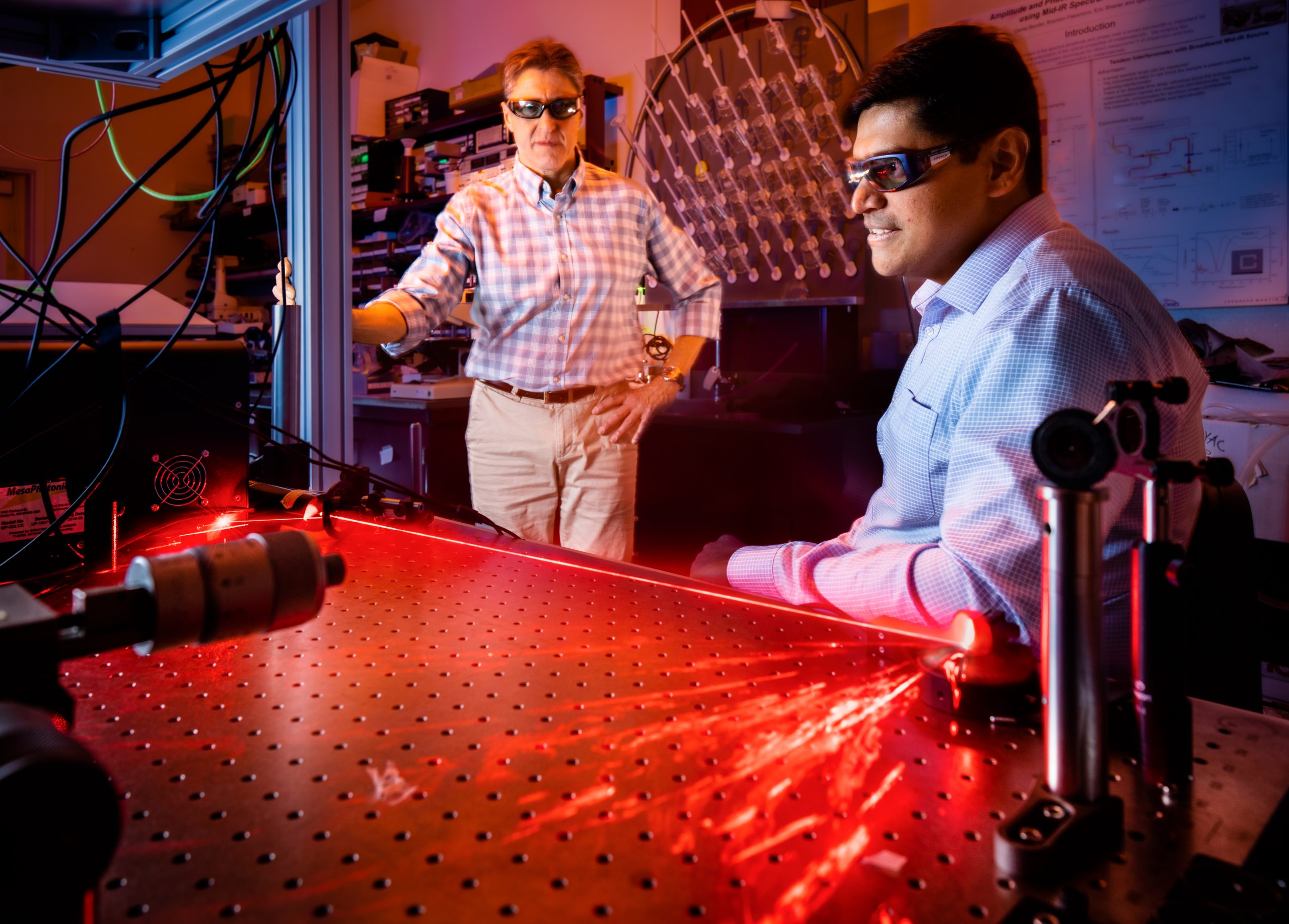 Two researchers stand adjacent to optical equipment emitting red light in a lab