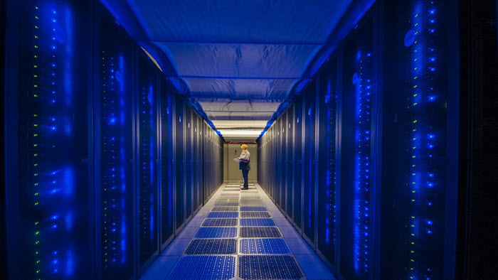 A scientist stands in the background amongst rows of supercomputing servers.
