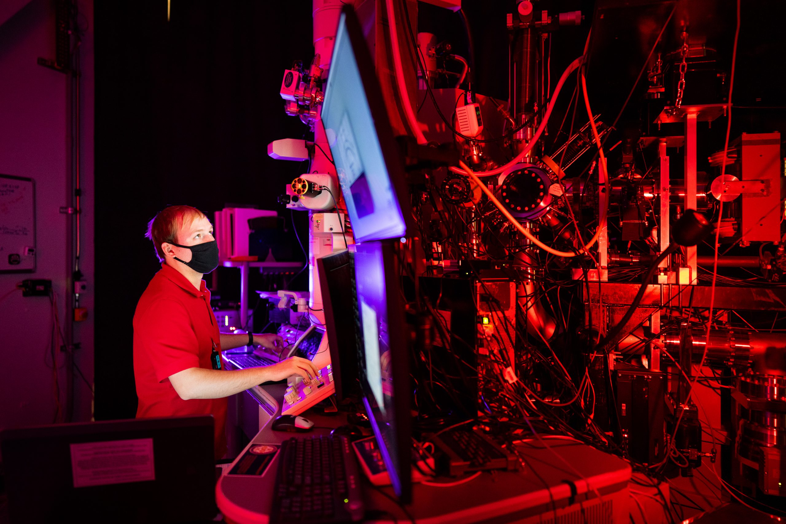 A researcher examines data on a large screen while standing among laboratory equipment lit with red light.