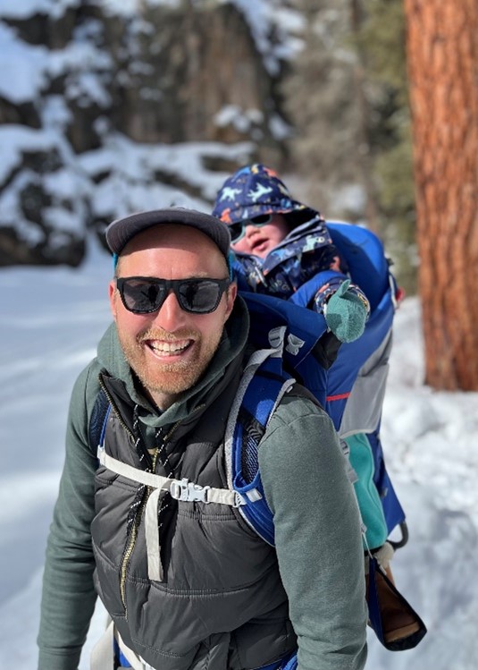 Andrew Knight smiling with a child in a backpack carrier in a snowy forest