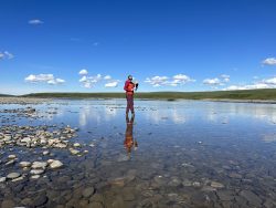 A researcher in a red jacket waves to the camera as they cross a shallow, pristine river in a verdant Arctic expanse.