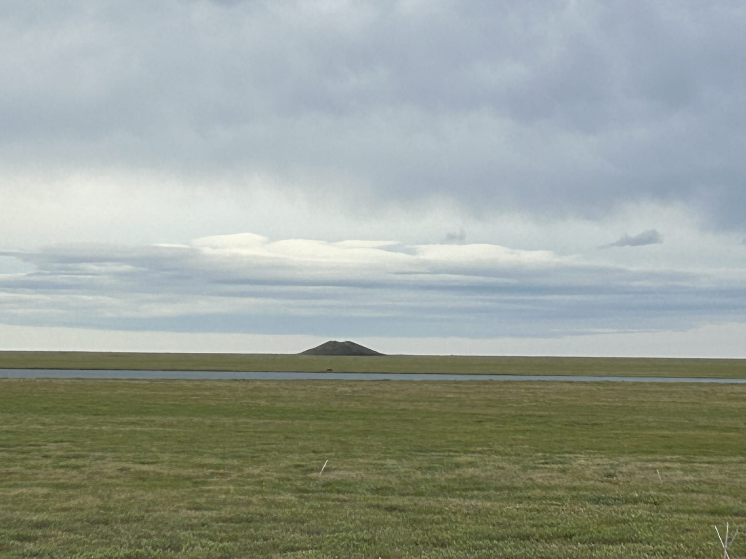 A verdant arctic tundra extends to the horizon. A small hill is apparent in the middle of the tundra -- it is a A permafrost-created pingo or “ice pimple” in the North Slope of Alaska.