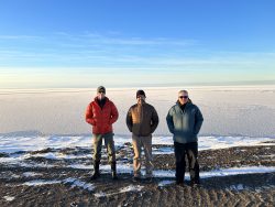 3 researchers stand in the forefront. An Arctic tundra extends behind them to the horizon.