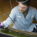 A researcher in a lab coat sits beside a flow way containing algae