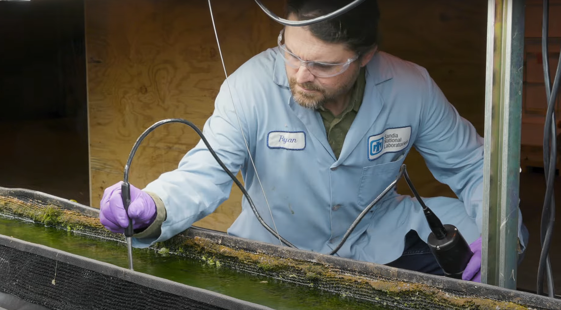 A researcher in a lab coat sits beside a flow way containing algae